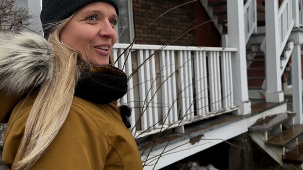 Colour photograph of Paule Vermot-Desroches, dressed in a yellow winter coat and a toque, in front of an apartment building balcony.