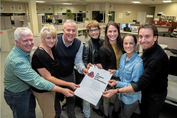 Photographie en couleur de sept personnes, 3 hommes et 4 femmes, dans une salle de rédaction et tenant une feuille de papier sur laquelle est écrit : « Je coopère pour Le Nouvelliste ».  