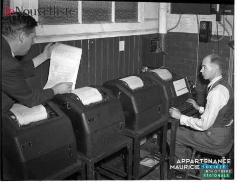 Photographie en noir et blanc de quatre machines à écrire et de deux hommes situés dans une pièce. Le premier est debout à gauche et vérifie un texte qui vient d’être produit par la machine. Le second est assis à droite et écrit sur le clavier de l’une des machines. Des appareils électriques sont posés sur le mur au fond de la pièce. 