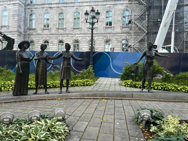 Photographie en couleur des statues en bronze de Marie Lacoste-Gérin-Lajoie, Idola St-Jean, Thérèse Forget-Casgrain et Marie-Claire Kirkland. Derrière eux se trouve un lampadaire et la devanture en brique de l’hôtel du Parlement du Québec. 