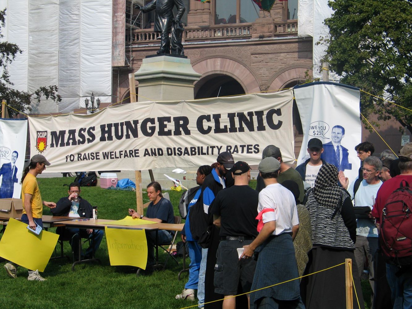 Une grande banderole portant l’inscription « Mass Hunger Clinic to Raise Welfare and Disability Rates » (Clinique de la faim massive pour augmenter les prestations sociales et les allocations d’invalidité) est déployée devant l'Assemblée législative de l'Ontario. Deux personnes sont assises à une table d'information, tandis que d'autres personnes font la file ou discutent.