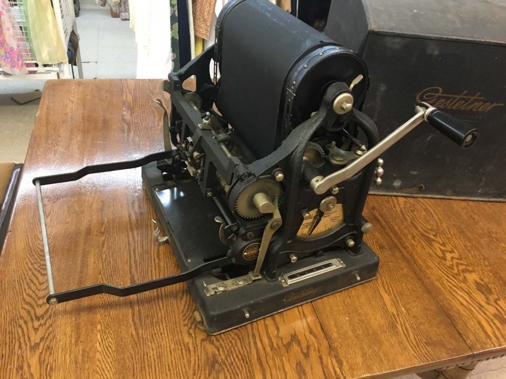 Une presse Gestetner noire posée sur une table en bois. Elle laisse apparaître ses mécanismes internes : rouleaux, engrenages, cadrans et une manivelle. En arrière-plan, on aperçoit un coffret noir sur lequel figure l'inscription « Gestetner » en lettres cursives.