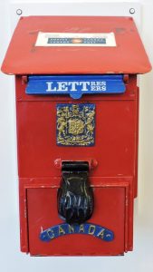 Une boîte aux lettres rouge en métal de style ancien, surmontée d'un logo plus moderne de Postes Canada. Sur le devant, on trouve un rabat bleu portant l'inscription « Letters » en anglais et « Lettres » en français. Sous le rabat, on trouve une gravure ornée représentant un emblème, un couvre-serrure noir et l'inscription « Canada ».