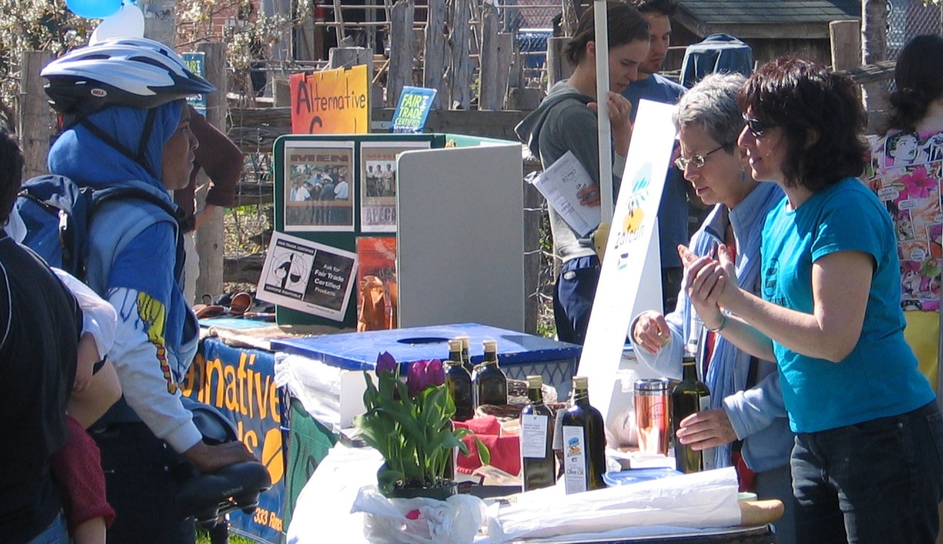 Une femme debout derrière la table de vente en plein air où sont exposées des bouteilles d'huile d'olive palestinienne, conversant avec une autre femme.