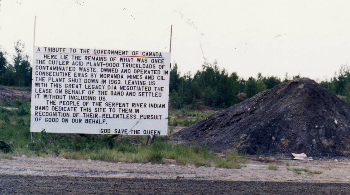 Un panneau situé au bord de l'autoroute, avec à sa droite, un amoncellement de déchets toxiques, sombres . Le panneau indique : « Hommage au gouvernement du Canada. Ici reposent les vestiges de ce qui fut autrefois l'usine d'acide Cutler : 9 000 camions remplis de déchets contaminés. Propriété de Noranda Mines et CIL, qui se sont succédé à l'exploitation, l'usine a fermé ses portes en 1963, nous laissant cet extraordinaire héritage. La DIA a négocié le bail au nom de la bande et a conclu l'accord sans nous consulter. Le peuple de la Bande indienne de Serpent River leur dédie ce site, en reconnaissance de leur quête inlassable du bien en notre nom. Que Dieu sauve la Reine. »