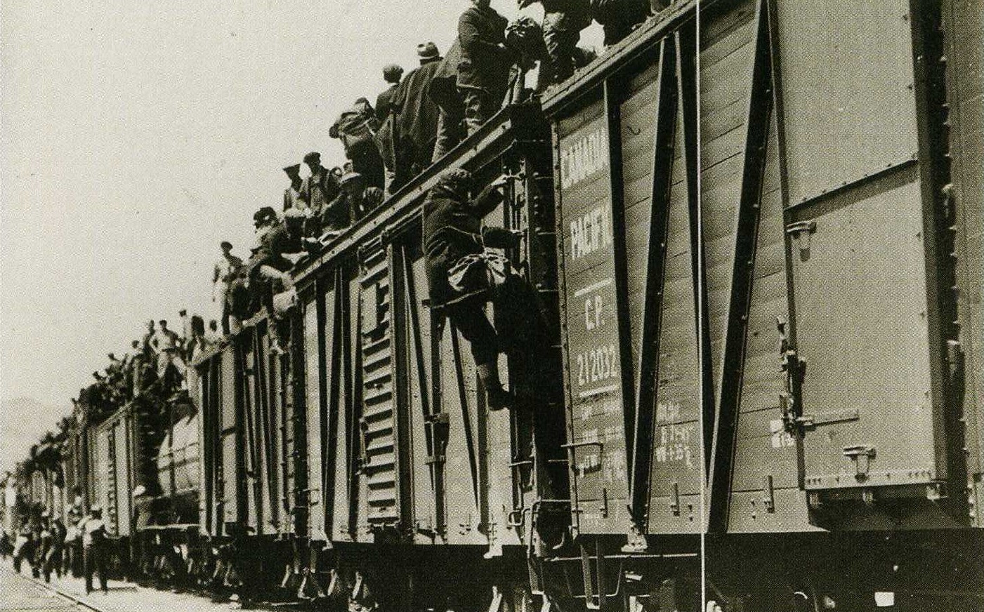 Photo en noir et blanc de travailleurs debout sur le toit d'un train du Canadien Pacifique, tandis que d'autres, debout près des rails, tentent de monter à bord. Le point focal de la photo montre un homme qui grimpe sur le train pour rejoindre le groupe debout sur le toit.