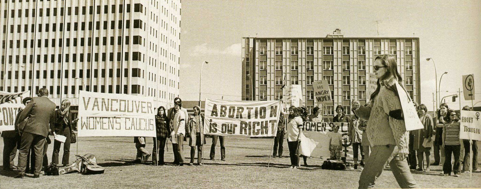 Photo en noir et blanc d'un groupe de femmes manifestantes debout devant deux bâtiments, tenant des pancartes et des banderoles, l'une proclamant « L'avortement est notre droit » et l'autre « Vancouver Women's Caucus ». Une femme marche devant le groupe.