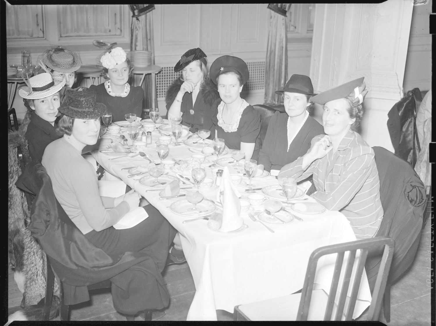 Photographie en noir et blanc présentant huit femmes, élégamment vêtues en tailleur et coiffées d'un chapeau, attablées autour d'une table dressée. 