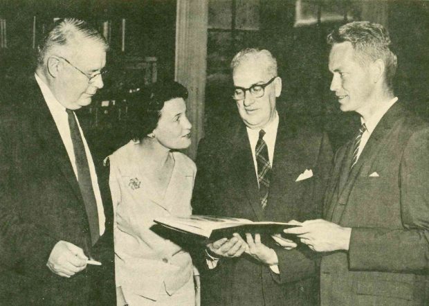 Photo en noir et blanc montrant trois hommes et une femme posant avec un livre.