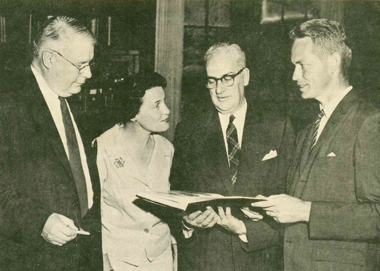 Photo en noir et blanc montrant trois hommes et une femme posant avec un livre.