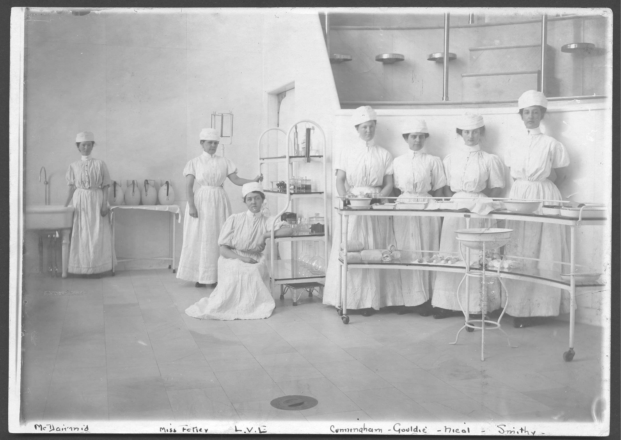 Photo en noir et blanc sur laquelle sept infirmières posent dans une salle d'opération, portant des uniformes d'infirmières du début du XIXe siècle.