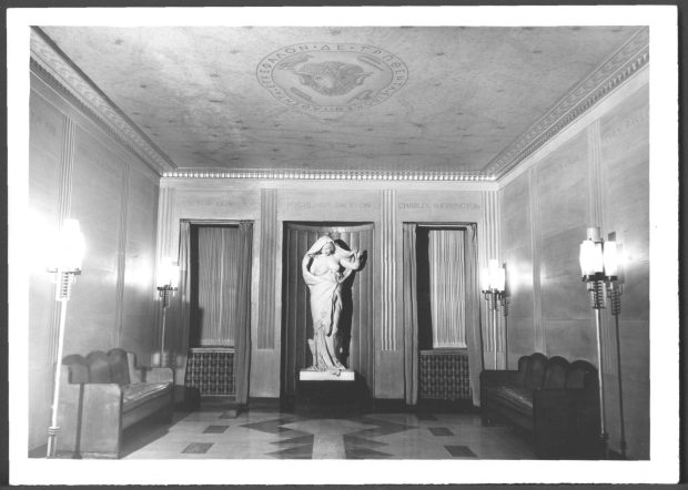 Image en noir et blanc d'un hall d'entrée carrelé contenant des bancs à droite et à gauche, des lustres et une statue en marbre blanc au centre.