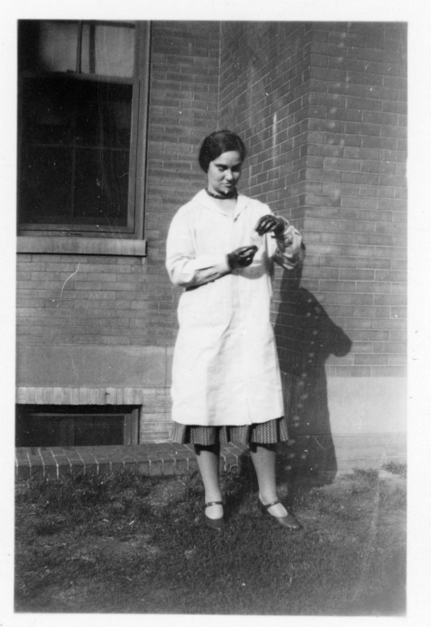 Photo en noir et blanc d'une femme portant une blouse blanche et des gants en caoutchouc, regardant un morceau de papier qu'elle tient entre ses mains.
