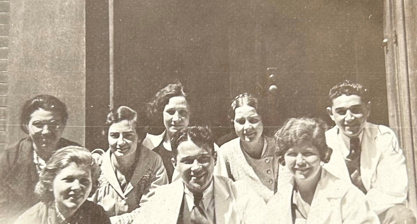Photo en noir et blanc d'un groupe d'élèves de tous genres portant des blouses de laboratoire et formant deux rangées. La photo est recadrée à hauteur d’épaule.   