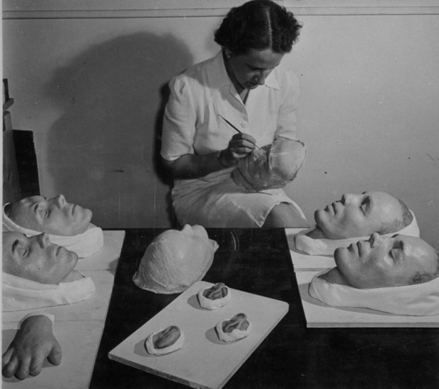 Photo en noir et blanc d'une femme peignant le moulage d'un visage. D'autres moulages reposent sur la table devant elle, dont des visages, une main et des oreilles.