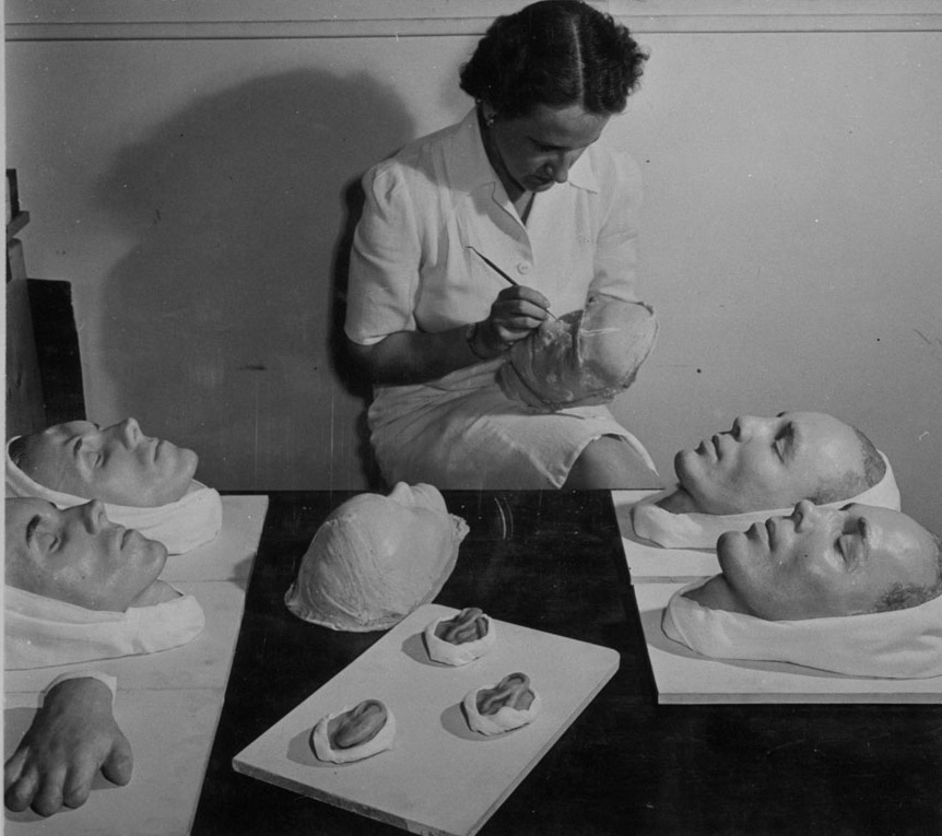 Photo en noir et blanc d'une femme peignant le moulage d'un visage. D'autres moulages reposent sur la table devant elle, dont des visages, une main et des oreilles.