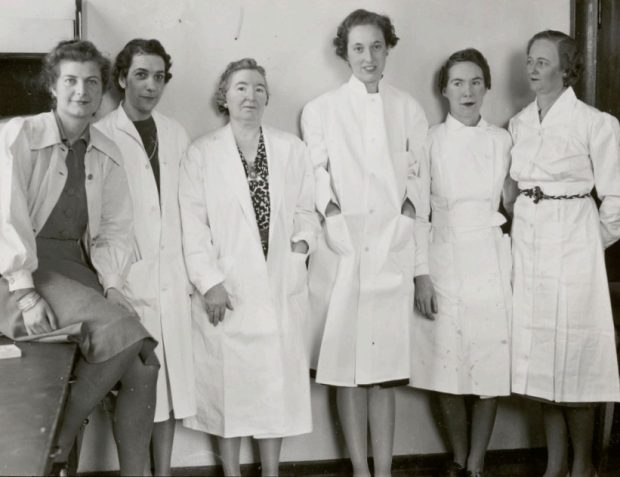Photo en noir et blanc de six femmes posant côte à côte, portant des blouses de laboratoire.