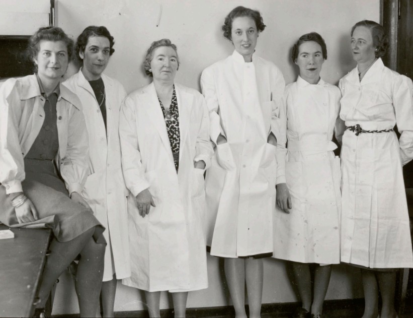 Photo en noir et blanc de six femmes posant côte à côte, portant des blouses de laboratoire. 