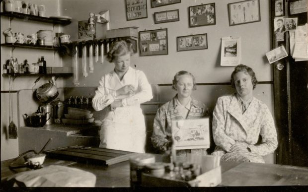Photo en noir et blanc de trois femmes assises dans un laboratoire. À gauche, un évier et une étagère où sont posés des tasses, une bouilloire et une théière. Le mur du fond est tapissé de portraits encadrés du personnel.
