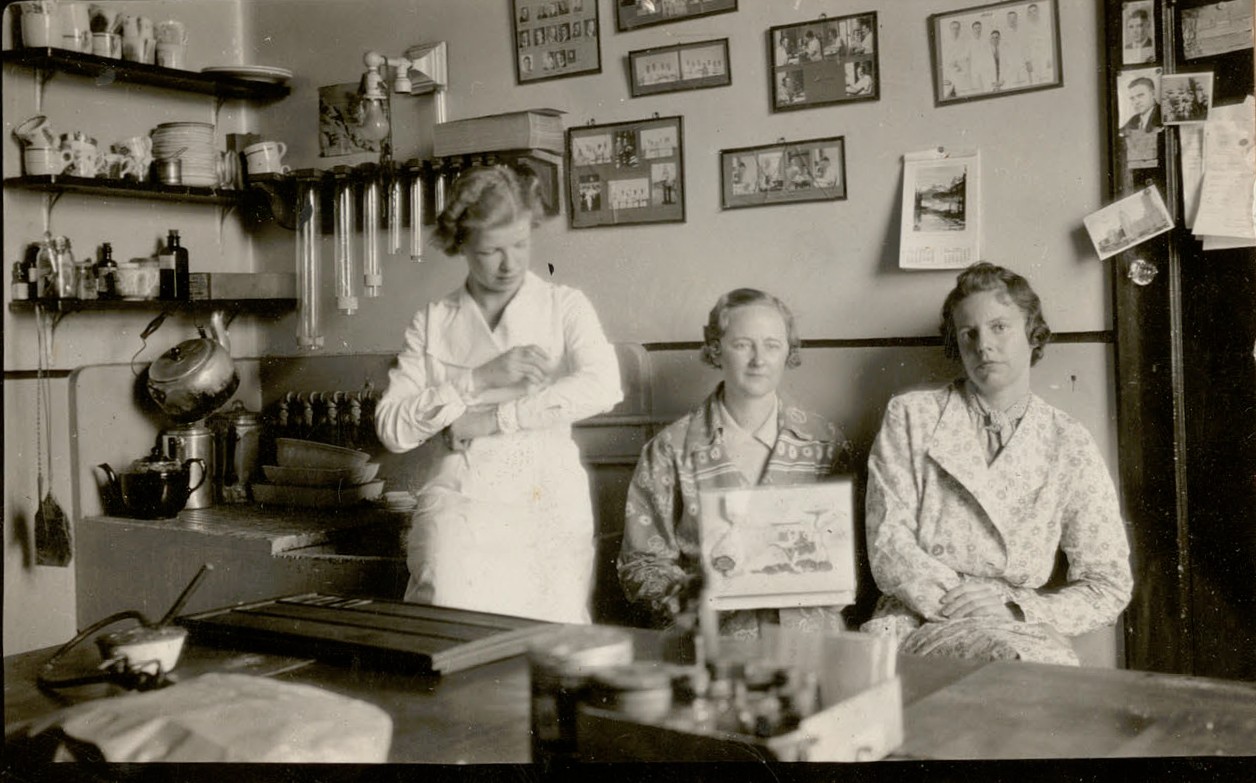 Photo en noir et blanc de trois femmes assises dans un laboratoire. À gauche, un évier et une étagère où sont posés des tasses, une bouilloire et une théière. Le mur du fond est tapissé de portraits encadrés du personnel. 