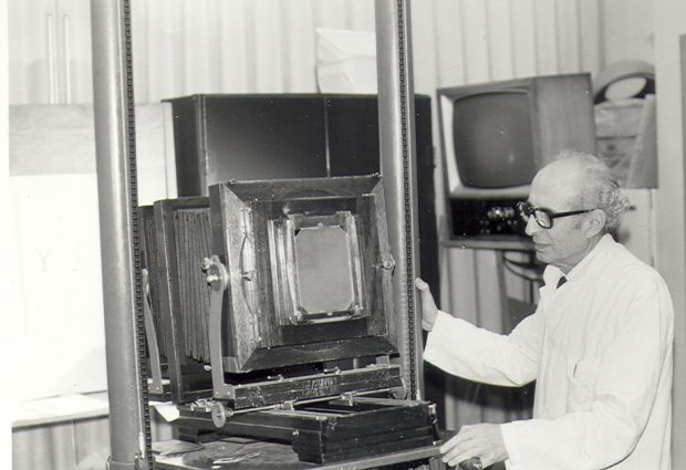 Photo en noir et blanc d'un photographe âgé maniant un grand appareil photo avec du matériel audiovisuel en arrière-plan.