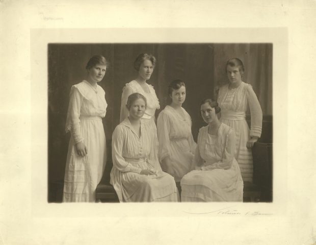 Photographie aux tons sépia montrant six femmes vêtues de robes blanches, posant et regardant l'appareil. Trois sont assises au centre, les trois autres debout.
