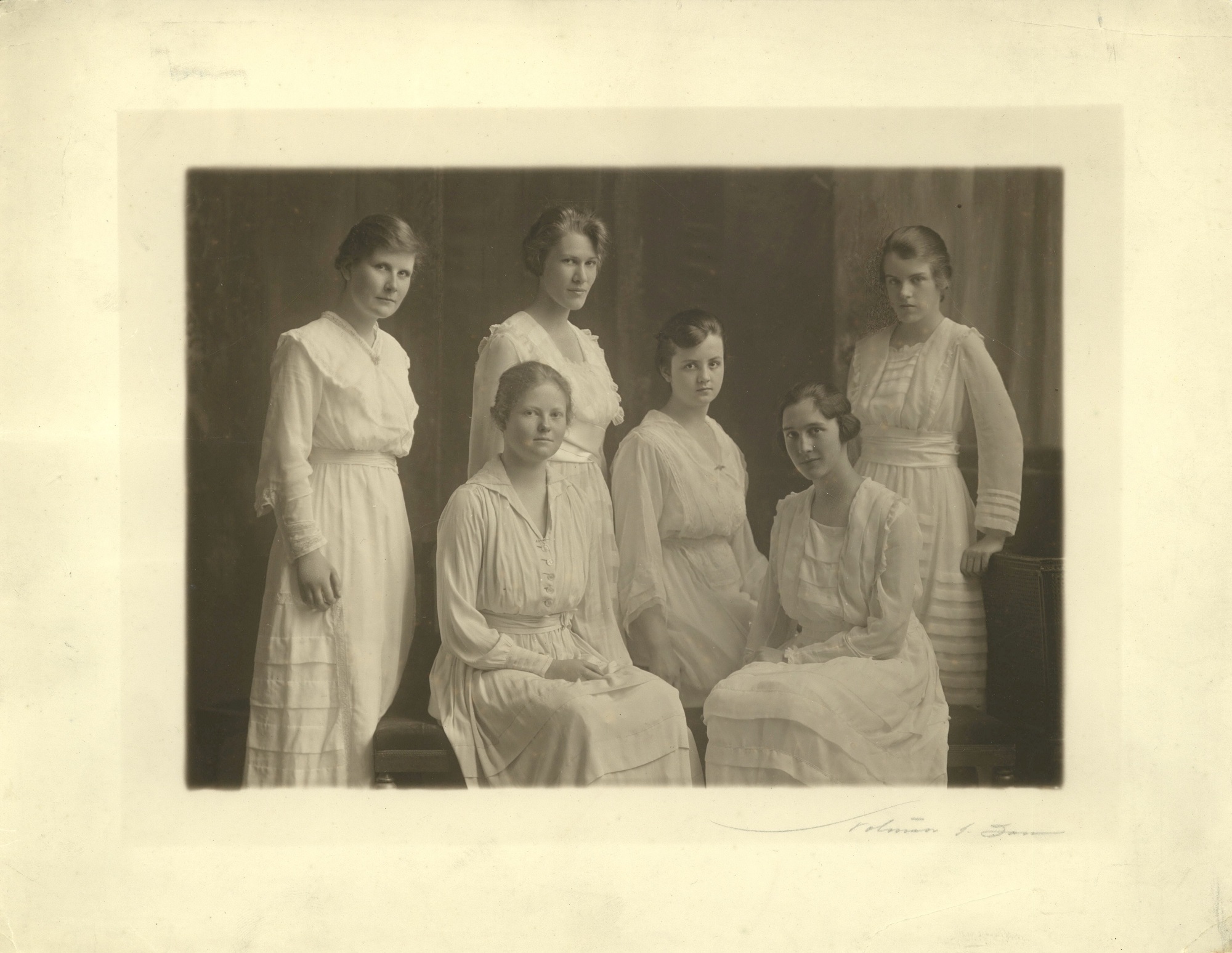 Photographie aux tons sépia montrant six femmes vêtues de robes blanches, posant et regardant l'appareil. Trois sont assises au centre, les trois autres debout. 