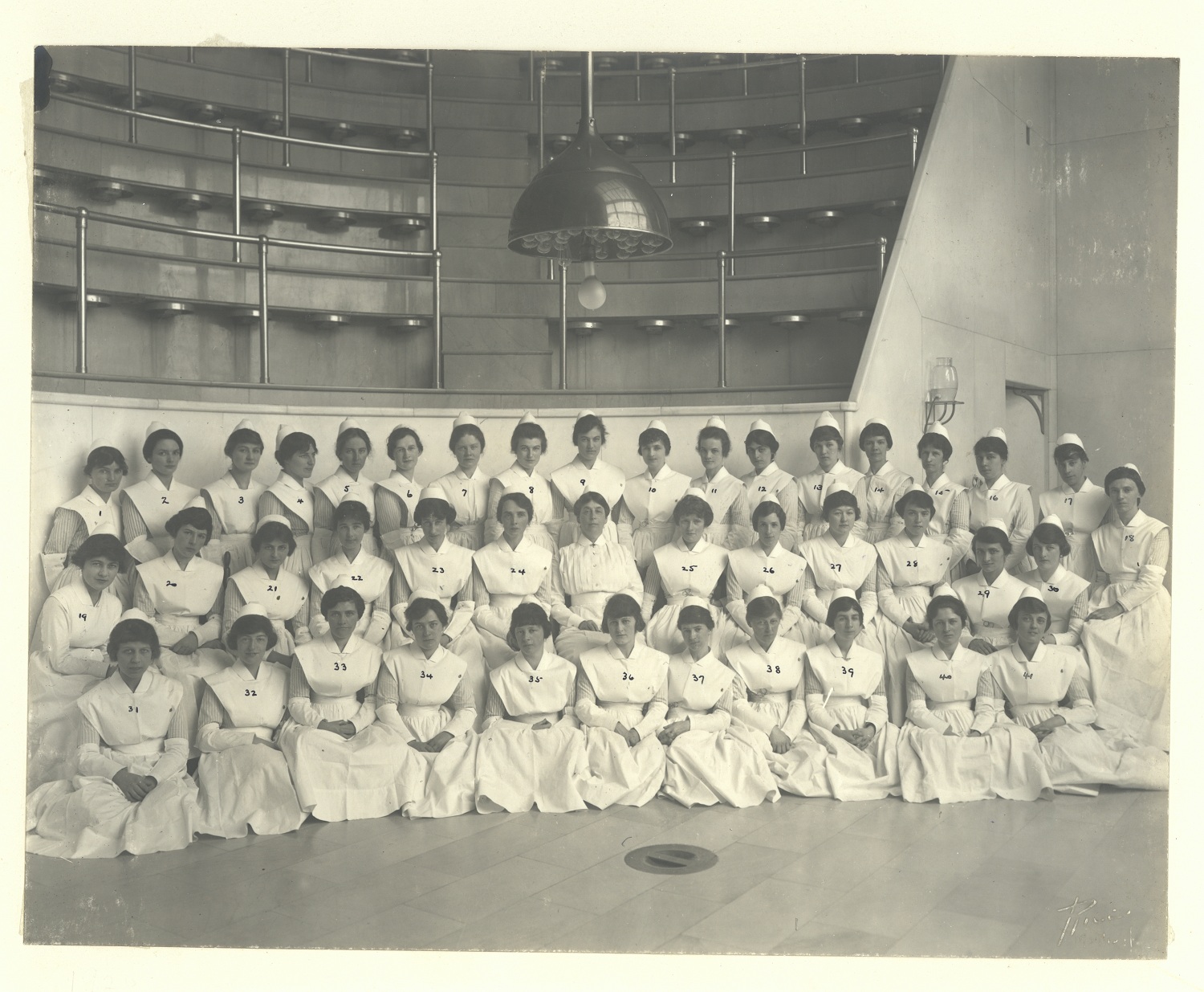 Photo en noir et blanc d'un groupe d'infirmières diplômées en uniforme blanc, posant dans une salle d'opération. 