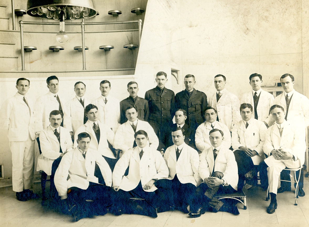 21 hommes posent en trois rangées dans l'amphithéâtre chirurgical d'un hôpital, vêtus de blouses blanches ou d'uniformes militaires. 