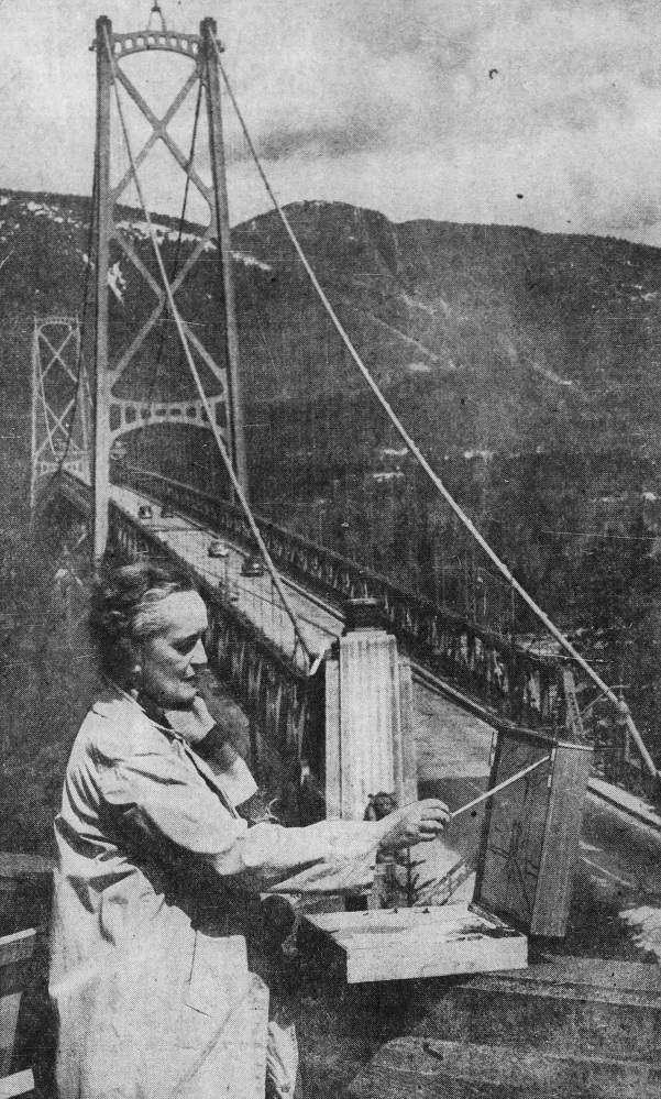 Photo en noir et blanc d'une femme peignant en plein air à l'entrée d'un pont suspendu, au milieu d'un paysage montagneux. 