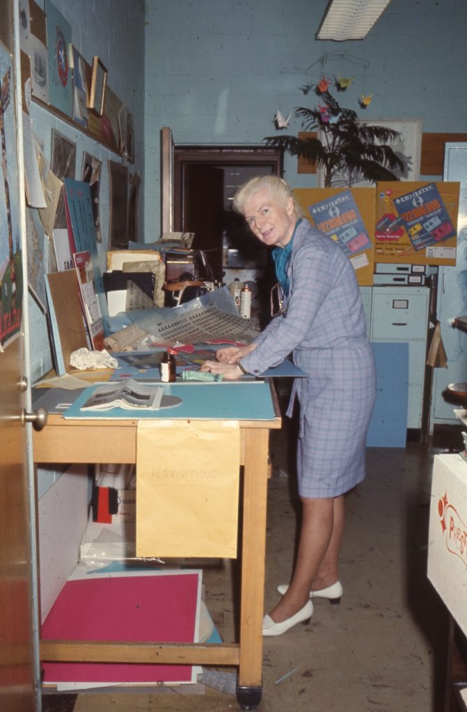 Photo en couleur de Mary Gzowski, penchée sur le bureau de son atelier, entourée de plusieurs projets et affiches sur son bureau et sur les murs.