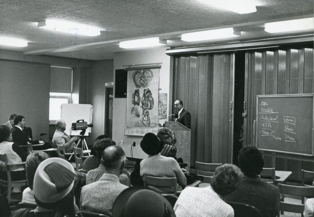 Photo en noir et blanc d'un médecin faisant une présentation à des personnes assises à des bureaux, à partir d’une illustration médicale de Pauline Larivière.