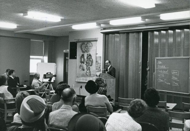 Photo en noir et blanc d'un médecin faisant une présentation à des personnes assises à des bureaux, à partir d’une illustration médicale de Pauline Larivière.