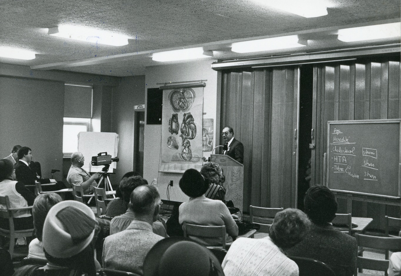 Photo en noir et blanc d'un médecin faisant une présentation à des personnes assises à des bureaux, à partir d’une illustration médicale de Pauline Larivière. 