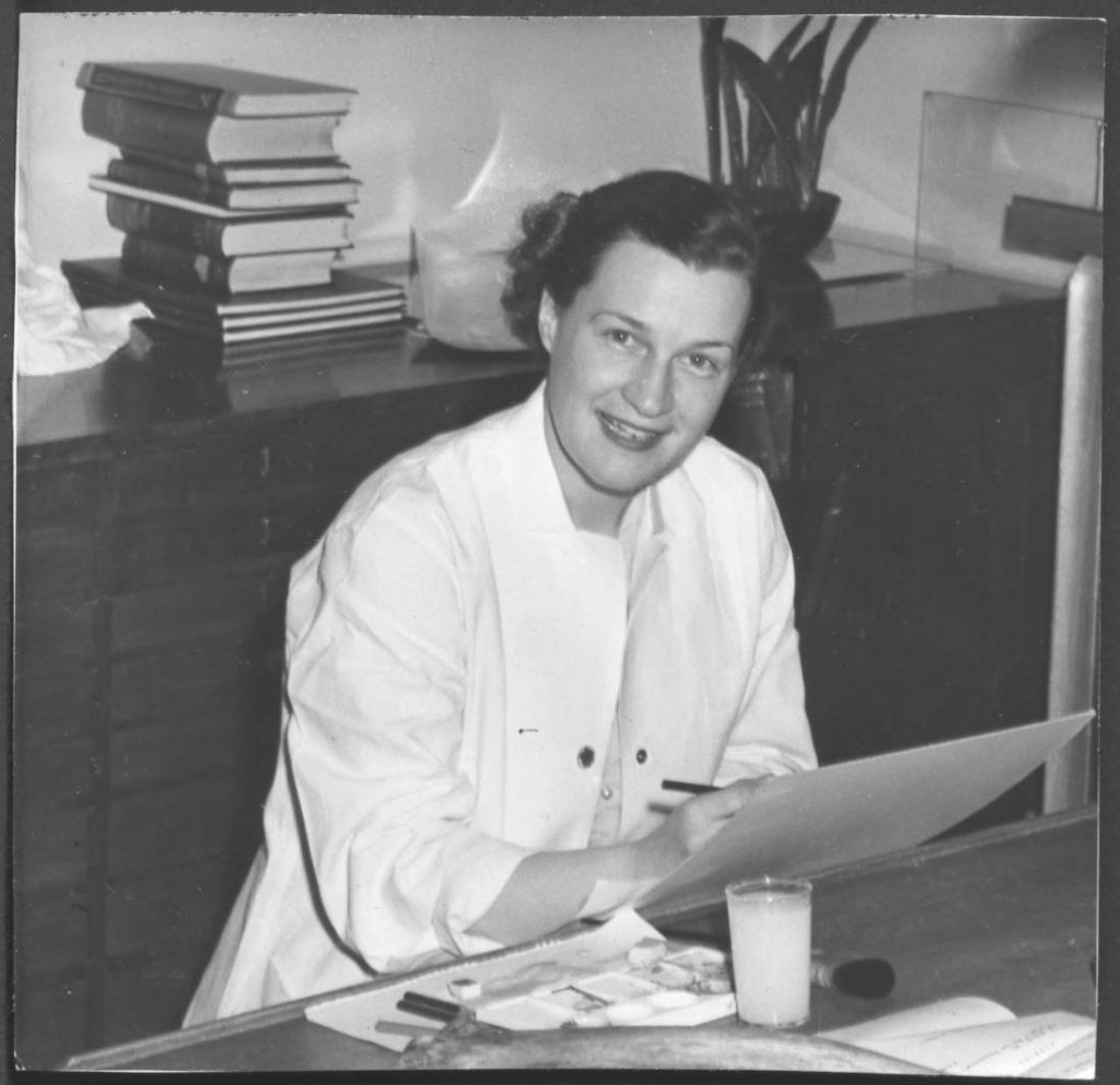 Photo en noir et blanc d'une femme assise à un bureau, dessinant sur une planche à illustration.