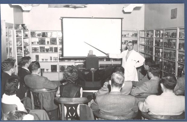 Photo en noir et blanc d'une salle de classe tapissée de diapositives et de spécimens pédagogiques médicaux. Faisant face à l'appareil photo, un homme en blouse blanche indique quelque chose aux élèves avec un pointeur.