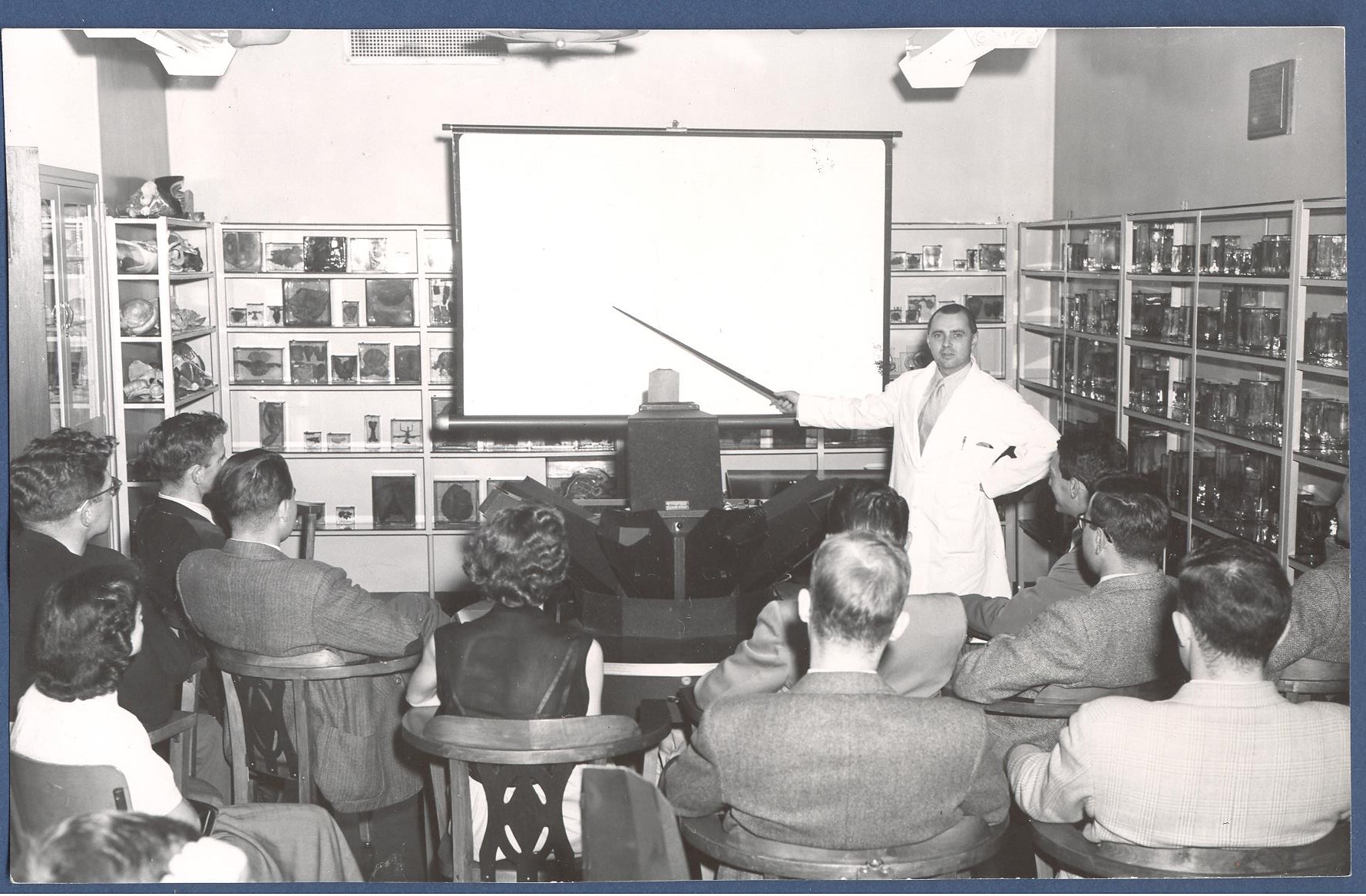 Photo en noir et blanc d'une salle de classe tapissée de diapositives et de spécimens pédagogiques médicaux. Faisant face à l'appareil photo, un homme en blouse blanche indique quelque chose aux élèves avec un pointeur. 