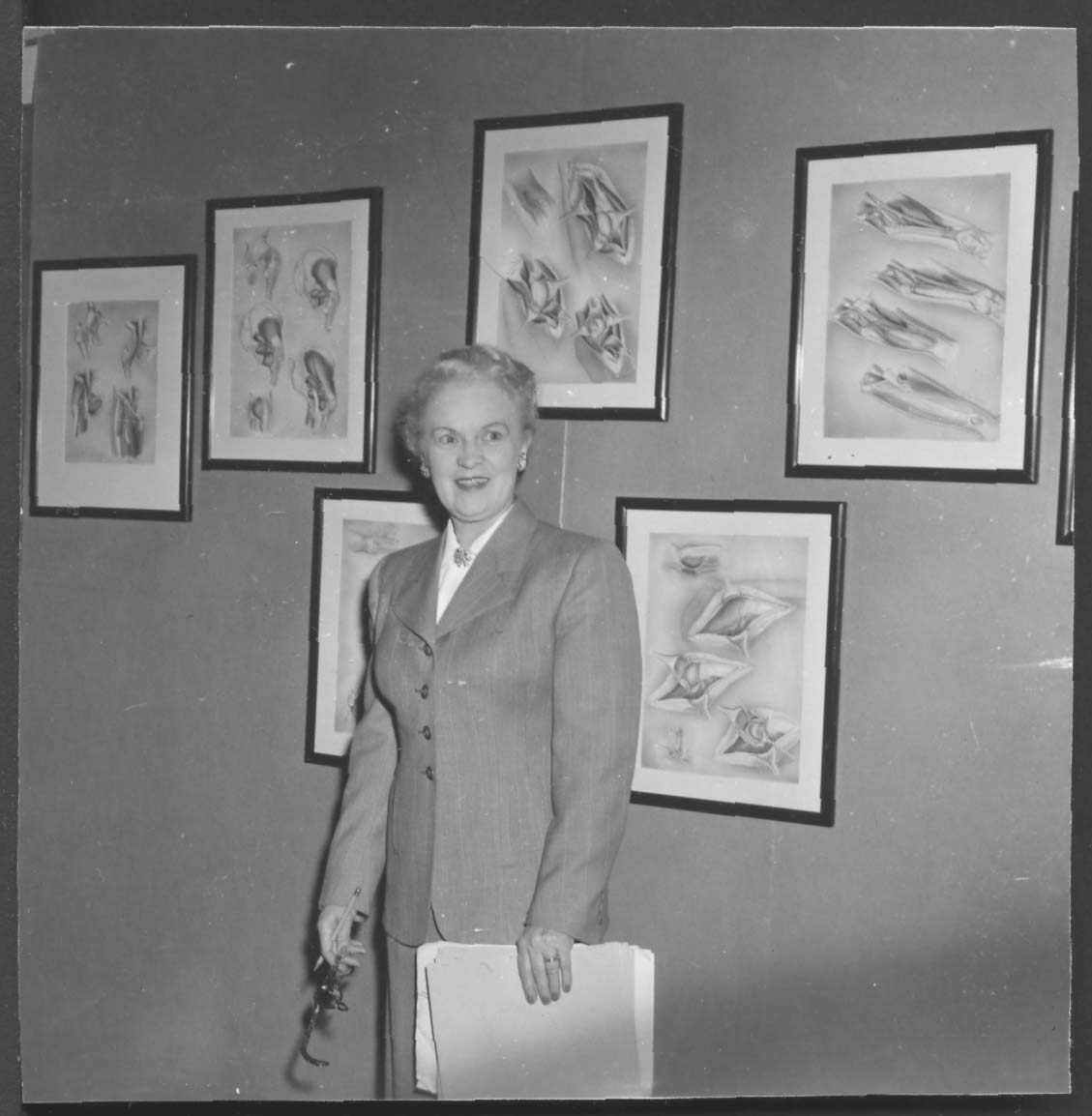 Photo en noir et blanc de Winifred Rock se tenant devant des illustrations d'Helen MacArthur. 