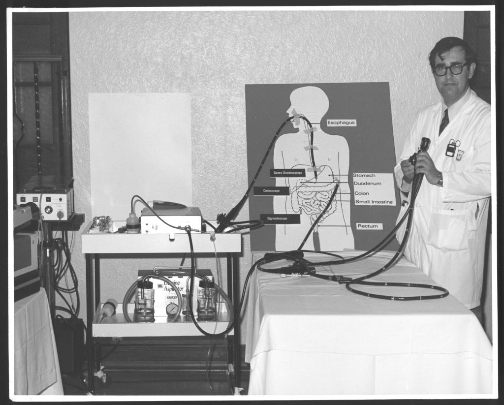 Photo en noir et blanc d'un médecin installant un dispositif d'enseignement médical comprenant une illustration du corps humain relié à divers tubes et équipements médicaux.