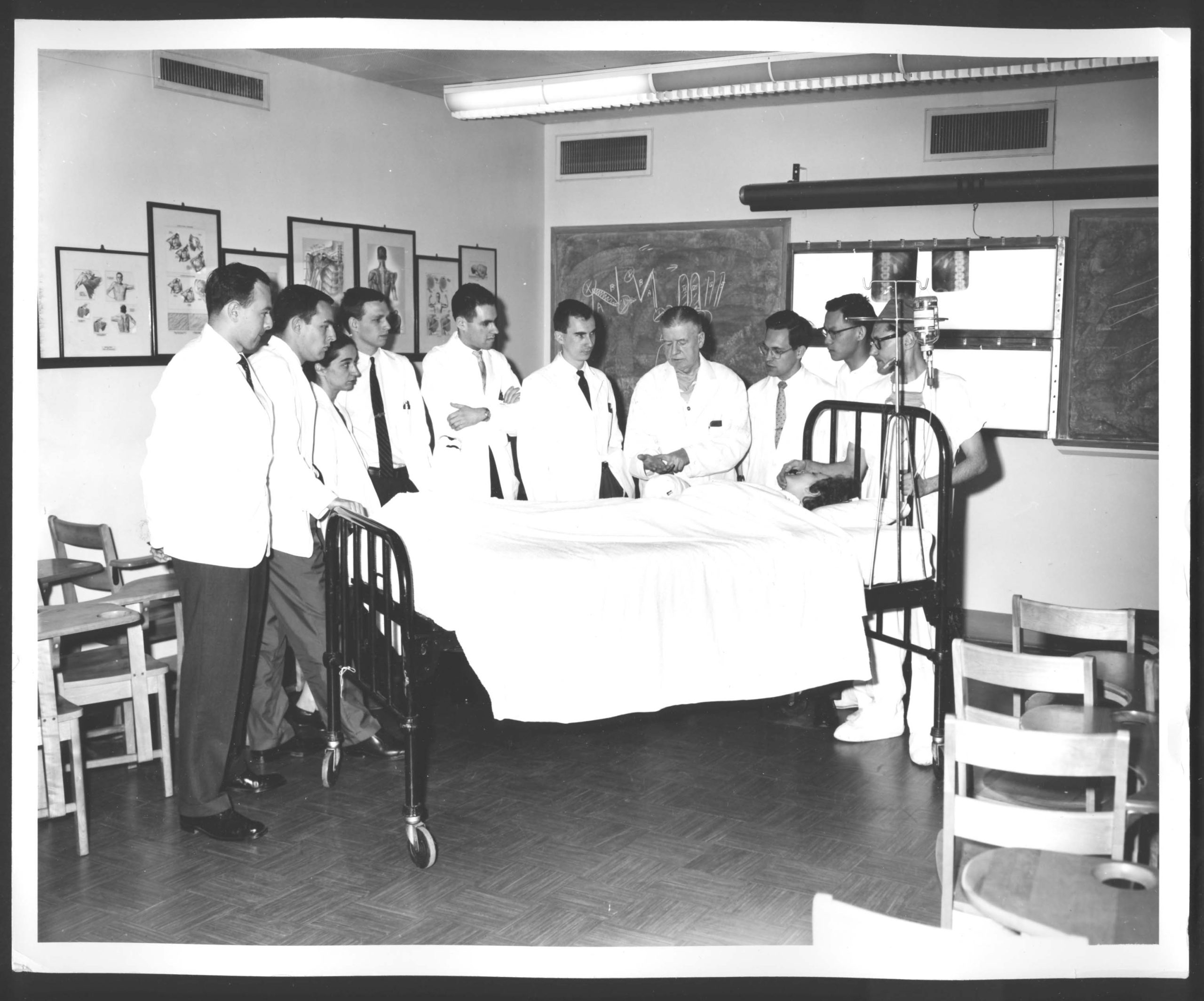 Photo en noir et blanc d'un médecin enseignant à des élèves résidents au chevet d'un patient. Des illustrations médicales sont accrochées en arrière-plan.