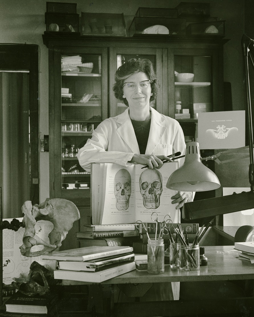 Photo en noir et blanc d’une femme portant une blouse de laboratoire dans un laboratoire, présentant un livre ouvert d’illustrations médicales. 