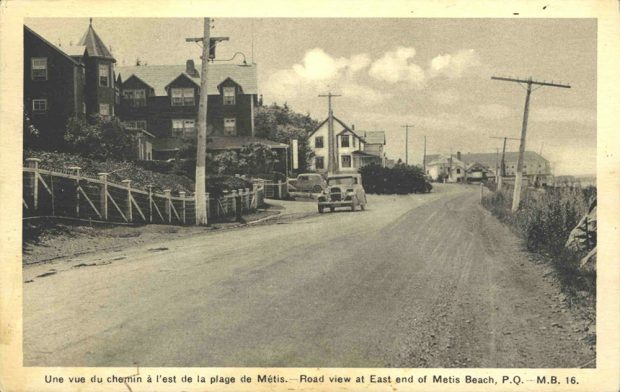 Carte postale noire et blanc imprimée sur carton présente le chemin du roi, la route de terre traversant la ville de Métis-sur-Mer. Une voiture est stationnée le long de la route. Le long de la route on y retrouve 2 des grands hôtels de l'époque ainsi que quelques résidences de villégiature. 