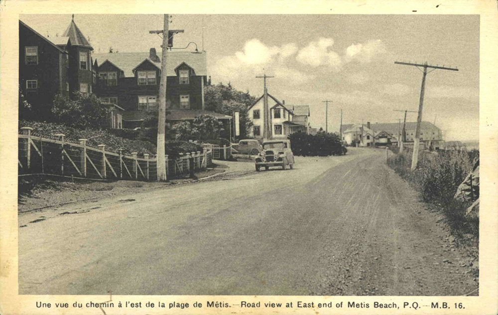 Carte postale noire et blanc imprimée sur carton présente le chemin du roi, la route de terre traversant la ville de Métis-sur-Mer. Une voiture est stationnée le long de la route. Le long de la route on y retrouve 2 des grands hôtels de l'époque ainsi que quelques résidences de villégiature.