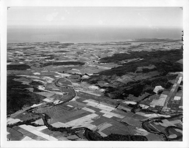 Photographie aérienne oblique noire et blanche montrant les terres agricoles et forestières longeant la rivière Mitis jusqu'au fleuve Saint-Laurent. Les différents types de culture se présentent comme un damier de couleurs et de textures différentes. 