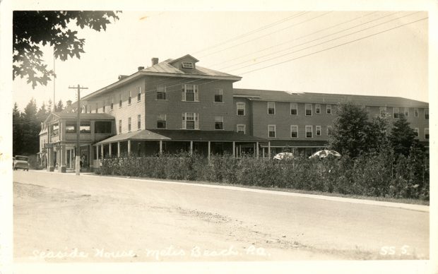 Carte postale présentant une photographie noir et blanc de l'hôtel Seaside de Métis-sur-Mer. L'imposant hôtel de bois à trois étages a deux ailes. L'une longe la route et l'autre s'allonge vers l'arrière à partir du centre de la première aile. La cour est cachée par une jeune haie de cèdres qui laisse paraître deux parasols.