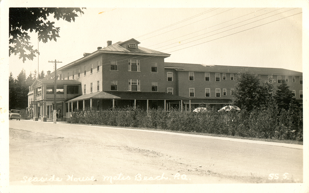 Carte postale présentant une photographie noir et blanc de l'hôtel Seaside de Métis-sur-Mer. L'imposant hôtel de bois à trois étages a deux ailes. L'une longe la route et l'autre s'allonge vers l'arrière à partir du centre de la première aile. La cour est cachée par une jeune haie de cèdres qui laisse paraître deux parasols.