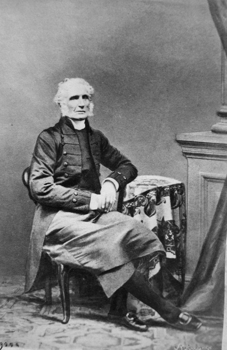 Portrait en studio en noir et blanc de l’évêque George Jehoshaphat Mountain. Le vieil homme est assis sur une chaise, vêtu des habits liturgiques d’un prêtre anglican. Le décor du studio évoque l’intérieur classique du style bourgeois de la seconde moitié du XIXe siècle.