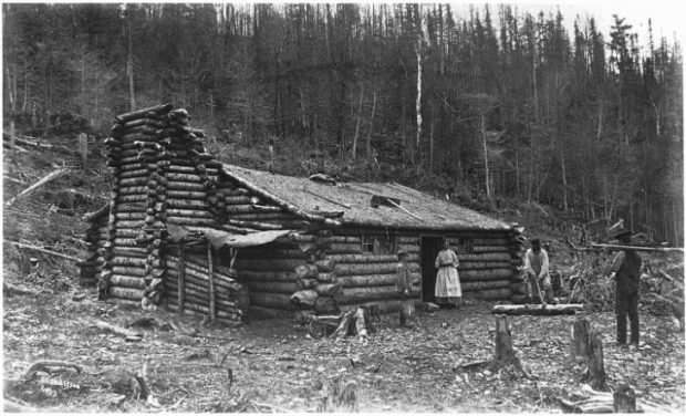 Photographie en noir et blanc d’une maison rustique en rondins. La maison est située au pied d’une colline et entourée de nombreuses souches d’arbres, probablement ceux qui ont servi à sa construction. Quatre personnes se trouvent à l’extérieur de la maison. Une femme en robe blanche se tient dans l’embrasure de la porte et nous regarde. À quelques pas d’elle, un jeune garçon tenant un seau en bois observe un homme en train de retirer l’écorce d’un tronc d’arbre posé au sol. Un autre homme s’approche de la scène, portant une charge sur ses épaules.