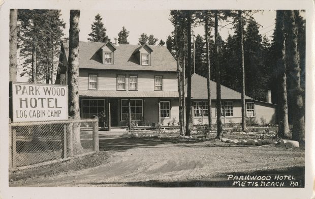 Carte postale présentant une photographie noir et blanc de l’entrée du Park Wood Hotel. L’hôtel est une grande maison de trois étages avec une annexe d’un étage. L’hôtel est entièrement fait de bois et est encerclé d’une forêt d’épinettes. Au premier plan de la photographie, on aperçoit le chemin d’accès à l’hôtel. Sur une clôture en grillage est disposée l’affiche de bois de l’hôtel, on peut y lire Park Wood Hotel Log Cabin Camp.