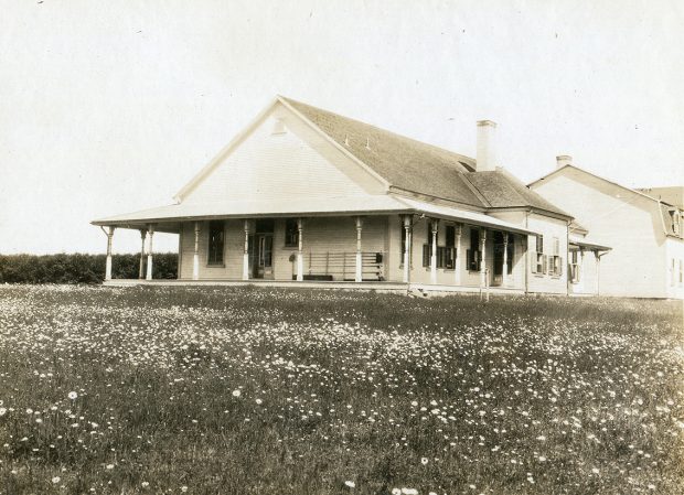 Photographie noir et blanc des façades nord et ouest de villa Estevan. L’extérieur du bâtiment est en déclin de bois et la toiture à double versant est faite de bardeau de cèdre. Les fenêtres sont munies de volets Le camp de pêche est muni d’une généreuse véranda sur laquelle est placée le support à canne a pêche. Au bout du bâtiment principal est raccordé au bâtiment secondaire occupé par les domestiques du propriétaire. Le bâtiment à 2 étages est légèrement reculé vers l’ouest par rapport au bâtiment principal. Le bâtiment est entouré d’un pré fleuri. L’on aperçoit l’immense haie de cèdre qui occupe l’espace avant du camp de pêche. Hormis sa taille, le camp de pêche est assez sobre, il n’y a que les colonnes du toit de la véranda qui souligne le style victorien de l’architecture du bâtiment. 